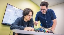 A student and instructor work together on a circuit board project, with a wiring diagram displayed on a screen behind them. The student focuses on the components, while the instructor gestures with a stylus. They are in a brightly lit classroom with light green walls.