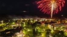 A nighttime aerial view of Saint Lawrence University’s campus lit with green trees and building lights, as red and gold fireworks burst in the sky above.