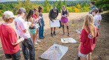 A group of Saint Lawrence University students stands in a circle outdoors, observing a peer drawing on a whiteboard with maps spread on the ground.