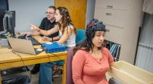 A student wearing a brainwave-monitoring cap sits with her arm in a water-filled container, while another student and a faculty member analyze data on a computer nearby.