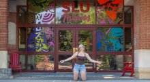 Student kneels and smiles in front of the Sullivan Student Center at Saint Lawrence University, with colorful window art and the words "SLU Thrives" above the entrance.