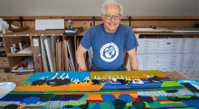 Woody Jackson, wearing a blue T-shirt stands behind a brightly colored painting featuring abstract shapes, bold stripes, and black-and-white cows in an art studio.